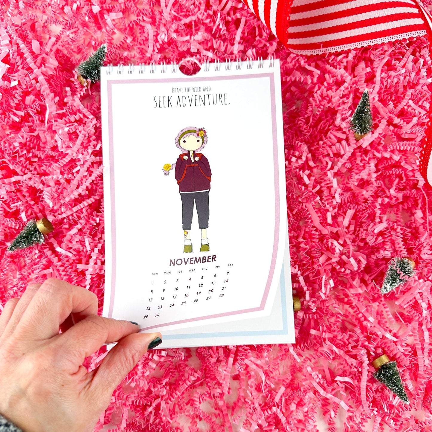 Hand holding a November calendar card with a person illustration on a pink textured background.